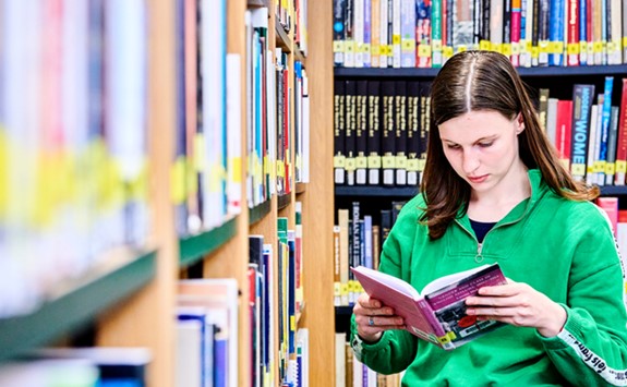Student standing and reading a book next to book shelves